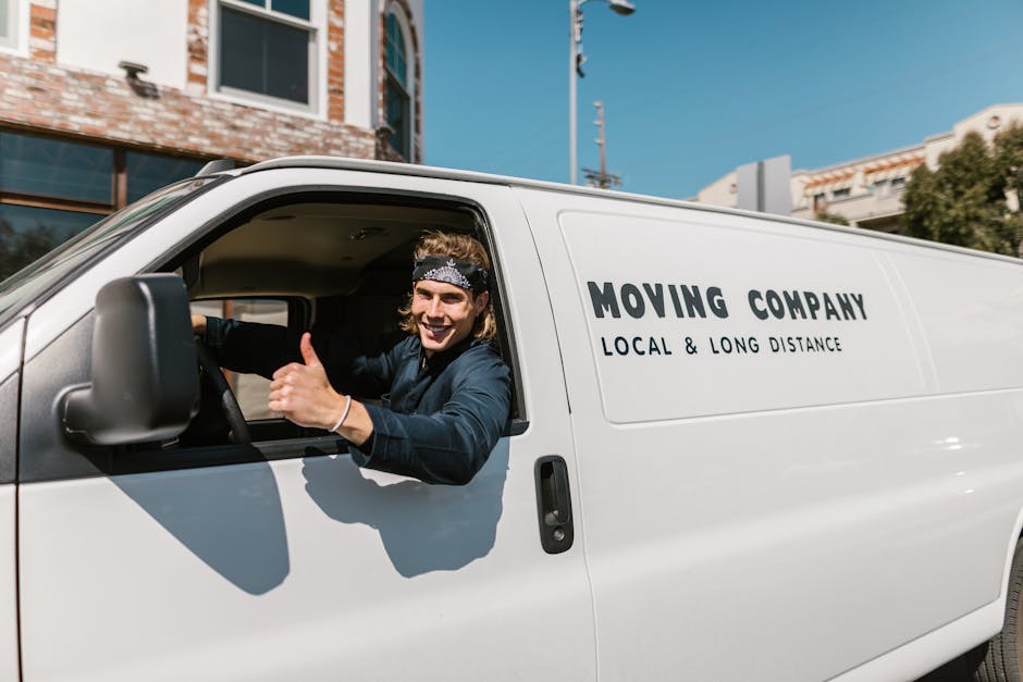 A male moving service worker with long hair and a patterned headband is sitting inside the cab of a white moving van, giving a thumbs-up. The van, branded with the logo 'MOVING COMPANY' and the phrase 'LOCAL & LONG DISTANCE,' is parked on a street with residential buildings and clear blue skies in the background. The interior of the vehicle shows part of the driver's seat and steering wheel, while the exterior indicates a professional home relocation or furniture transport operation, typical of house removals services provided by Colliers Wood Man and Van. The scene suggests a successful loading or moving process, with the van ready to assist with packing and moving tasks involving boxes, furniture, and equipment.