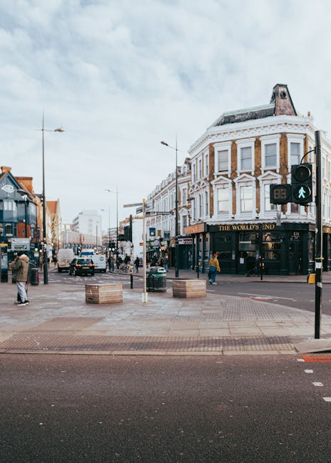 A view of a busy street corner in an urban area during daytime, featuring a historic three-storey building with white decorative trim and large windows, housing a pub named 'The World's End' at ground level. The sidewalk in front of the building is occupied by pedestrians walking and standing, with some waiting at a traffic light and others carrying small parcels or backpacks. Several vehicles, including cars and a delivery van, are visible on the street and parked along the curb. In the foreground, a trolley or dolly with boxes and packing materials may be present, indicating a home relocation or moving process. The scene is well-lit with natural daylight, and there are street lamps and signage, including a green pedestrian crossing signal. As part of professional removals services in Colliers Wood, Colliers Wood Man and Van might operate in this area to assist with furniture transport, packing, and logistics, supporting local house moves and urban relocations.