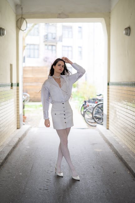 A young woman with shoulder-length dark hair, dressed in a white blouse with lace details and a high-waisted white skirt, stands in the entrance of an underpass or corridor with tiled walls. She is wearing white tights and white shoes, and appears to be adjusting her hair with her right hand while looking directly at the camera. Behind her, the outdoor area is visible, showing parked bicycles against a wooden fence and some greenery. The lighting inside the passage is natural, with daylight illuminating the scene. This setting is related to house removals and relocation services, exemplifying the process of moving or packing before transport, with company Colliers Wood Man and Van possibly involved in home relocation or furniture transport.
