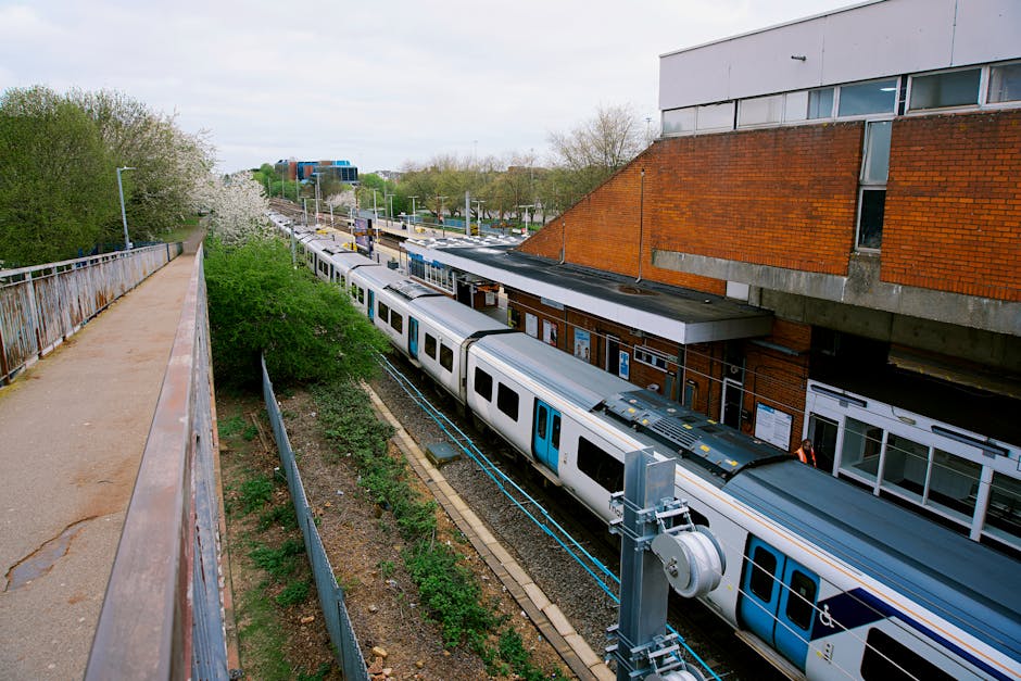 A view from an elevated walkway overlooking a train station platform where a silver and blue passenger train is stationary, with doors open for boarding or unloading. The platform features a brick building with signage and large windows, and a narrow perimeter area with a metal railing and small green shrubs separating it from the train tracks. Adjacent to the platform, a concrete extended structure supports upper floors of a building. In the background, trees with spring foliage and a clear sky are visible. This scene relates to the logistics of house removals and furniture transport, illustrating the loading and unloading environment typically involved in home relocation and moving services offered by Colliers Wood Man and Van.