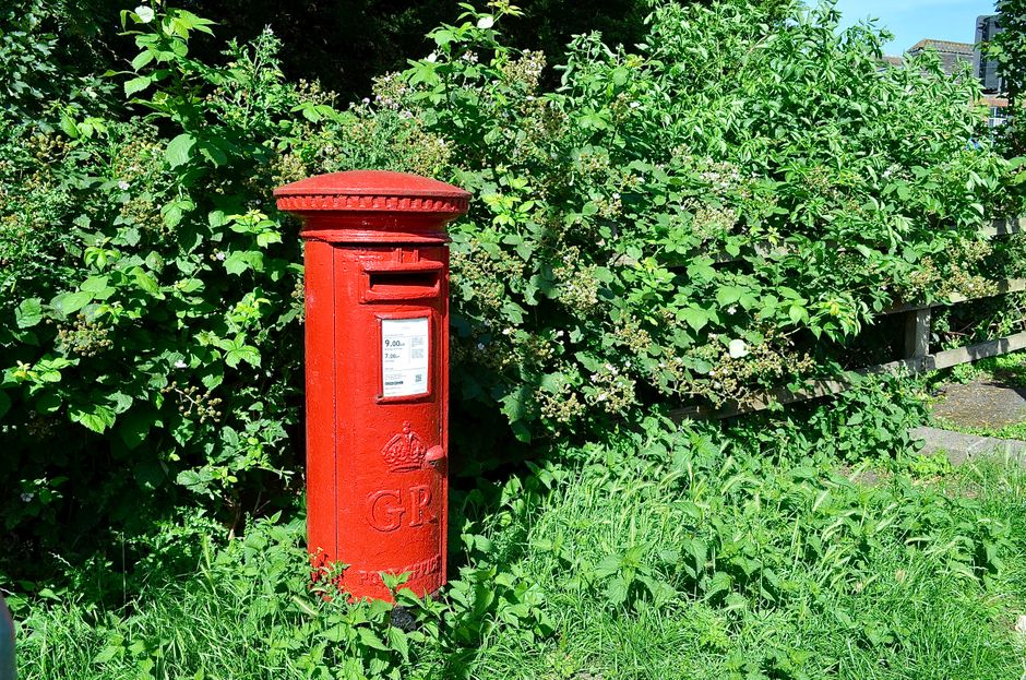 A red British pillar post box standing on a grassy verge beside a sidewalk, with a background of dense green foliage including bushes and trees. The post box has a small rectangular slot for mail, a white informational decal displaying collection times, and the letters 'GR' embossed on the lower part. The scene is outdoors in natural daylight, and the post box is part of a typical residential or suburban street setting, reflecting the urban environment where house removals and furniture transport services by Colliers Wood Man and Van are often required.